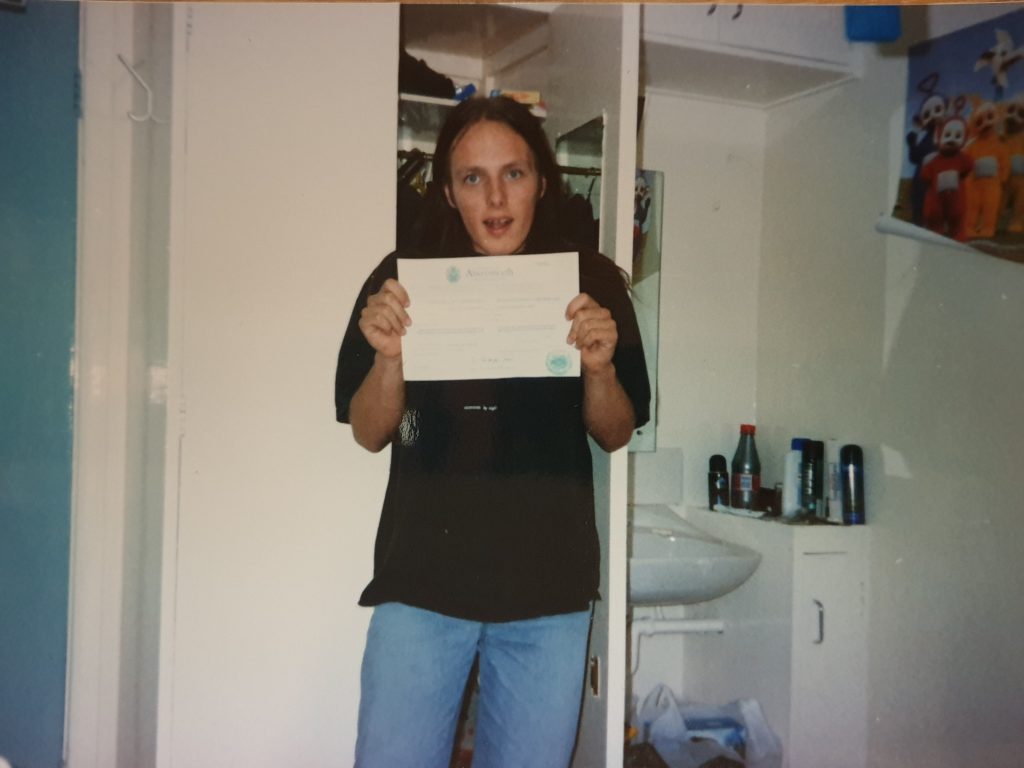 Standing in a study bedroom, Dan shows off his certificate of admission to the University of Wales, Aberystwyth.