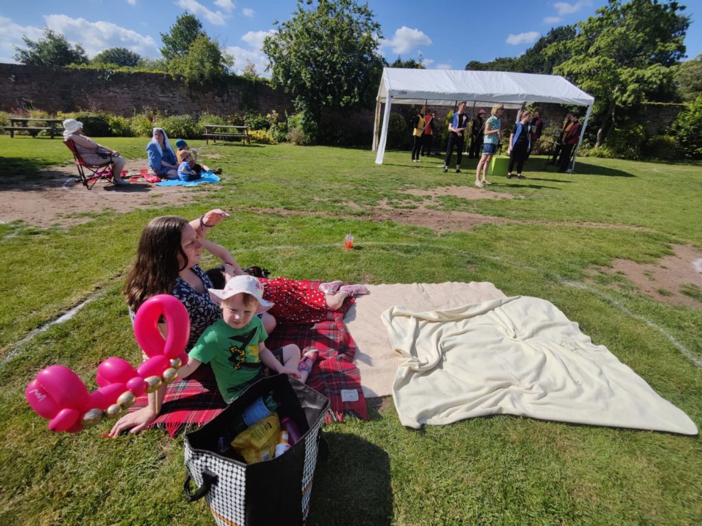 Ruth and John sit on a picnic blanket in a painted circle; the maquee for the band is behind them.
