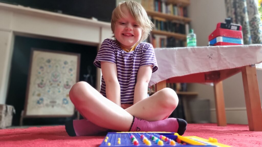 A young boy sits cross-legged on the floor, grinning excitedly at a Mastermind board (from the code-maker's side).
