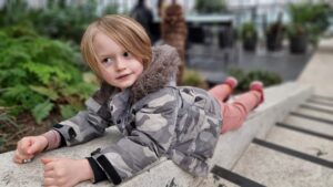 A 6 year old boy in a grey camouflage coat lies on a concrete wall alongside a staircase.