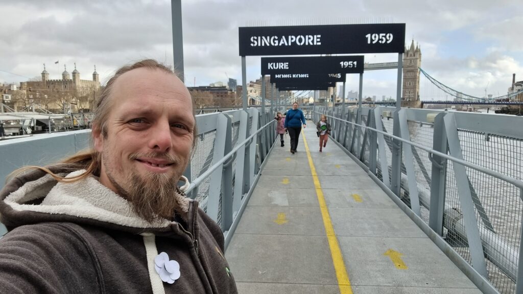 Dan stands on a pier leading out to the HMS Belfast, beneath a series of signs indicating where it served in particular years. In the distance, Ruth and the kids approach.