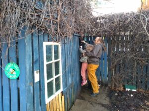Alongside a blue fence, Dan lifts up a small boy so he can reach a wall-mounted geocache.