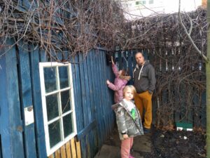 Dan stands with two children - a boy and a girl - near a high wall-mounted geocache; the girl is stretching to reach the cache container.
