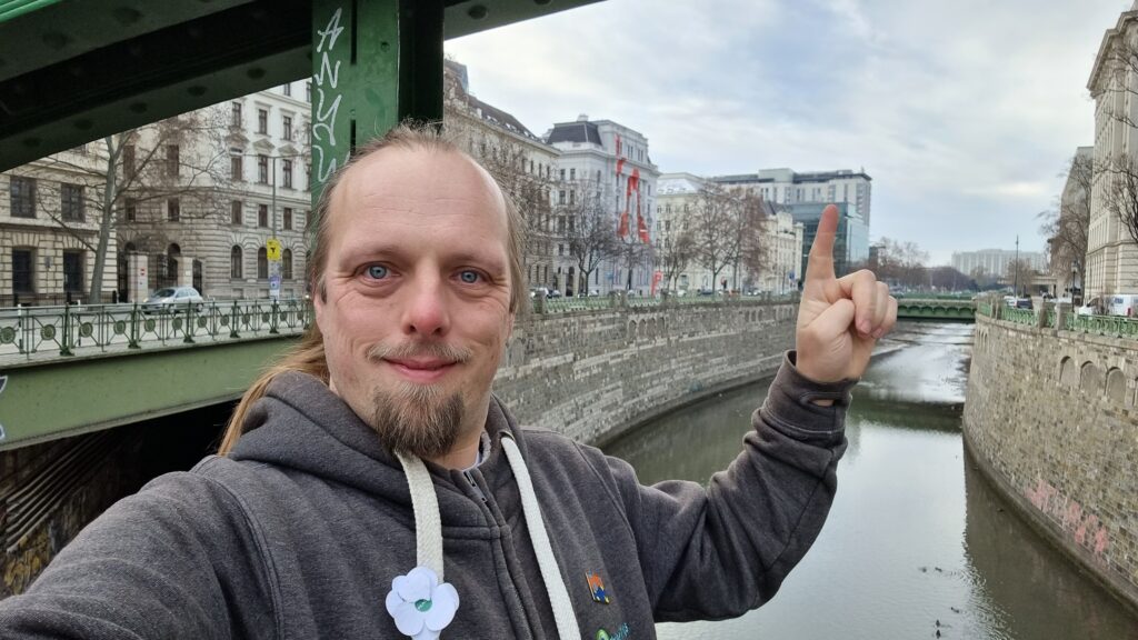 Dan stands on a bridge over the River Wein (before it meets the Danube), pointing to a window in the distance of a hotel.