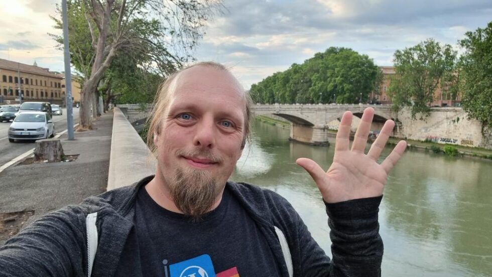Alongside the River Tiber, with a wide-arched stone bridge in the background, Dan sits on a wall and waves to the camera.
