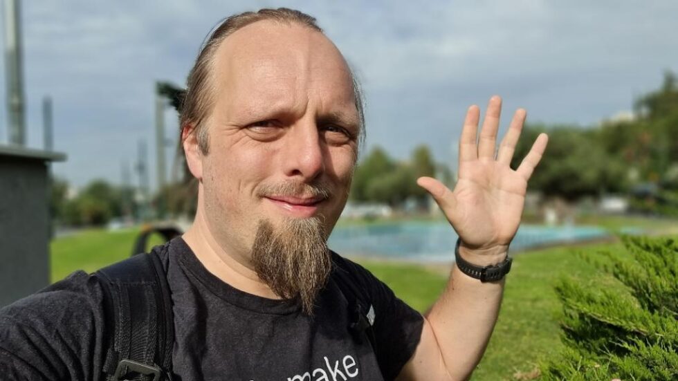 Dan, in a green park with water fountains, waves at the camera.