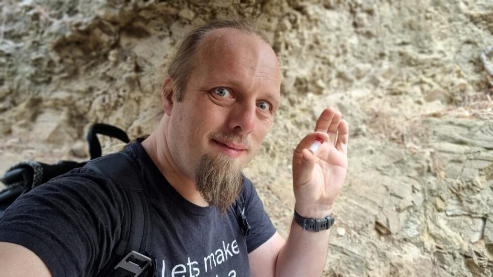 Dan, wearing a black t-shirt and a backpack, holds a tiny plastic container with a red lid. Behind him is a rough rock wall.