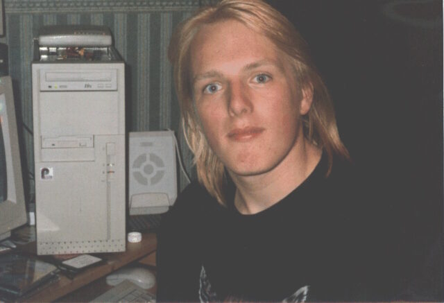 Photograph of Dan in his mid-teens, with shoulder-length bleached-blonde hair and wearing a t-shirt with a picture of a snarling wolf, sits in front of a running PC (with its beige case open) on which an external modem is precariously balanced.