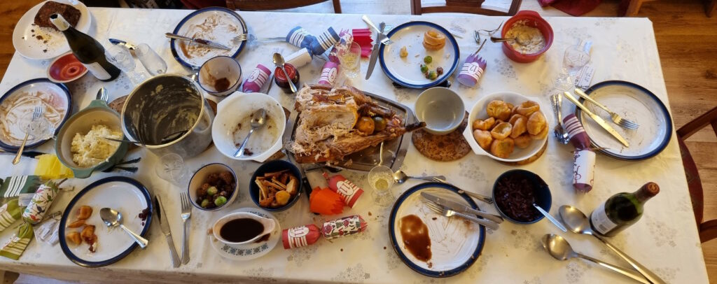 Top-down view of a dining table set with a Christmas-themed tablecloth. The meal has concluded and the seats have been vacated, but large amounts of food - most of a turkey, half a nutloaf, lots of mashed potato, several sprouts, stuffing balls, and chestnuts, some roast potatoes and parsnips, an entire boat full of gravy, and almost a dozen Yorkshire puddings - are still set out.