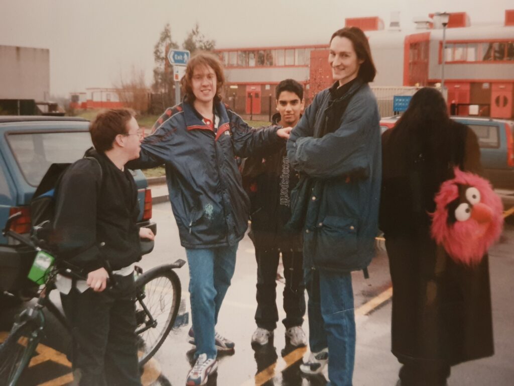 A group of young adults mill around in a rainy car park between campus buildings.