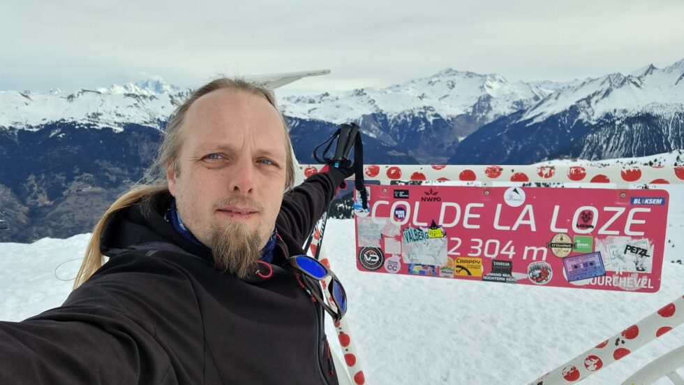 Dan, in a wintery mountainous landscape, poses for a selfie by a sculpture of a bicycle marking part of the route of the Tour de France.