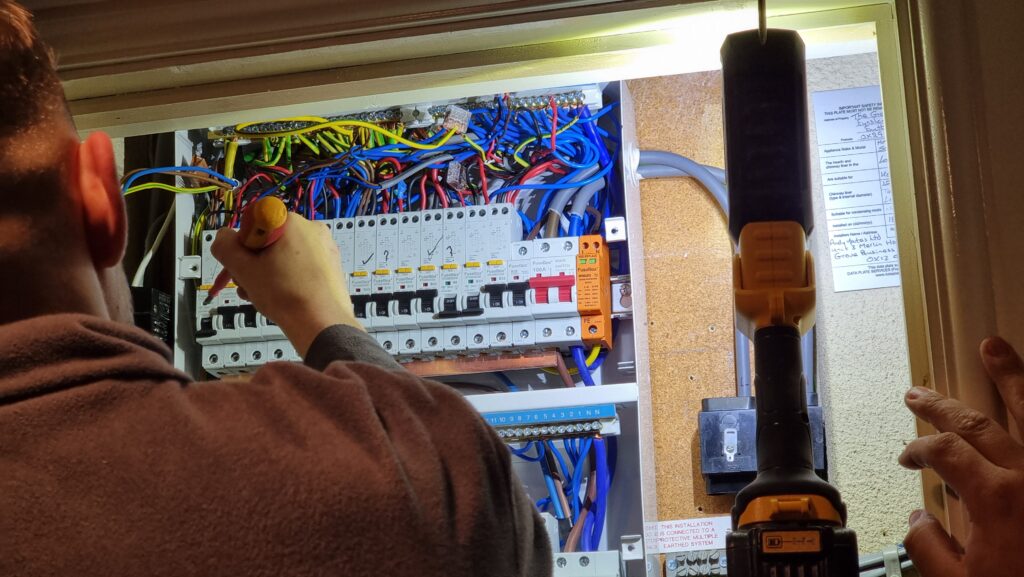 Electricity shining a torch into a cupboard containing an fusebox with an open cover; the switches are in a mixture of on and off positions.