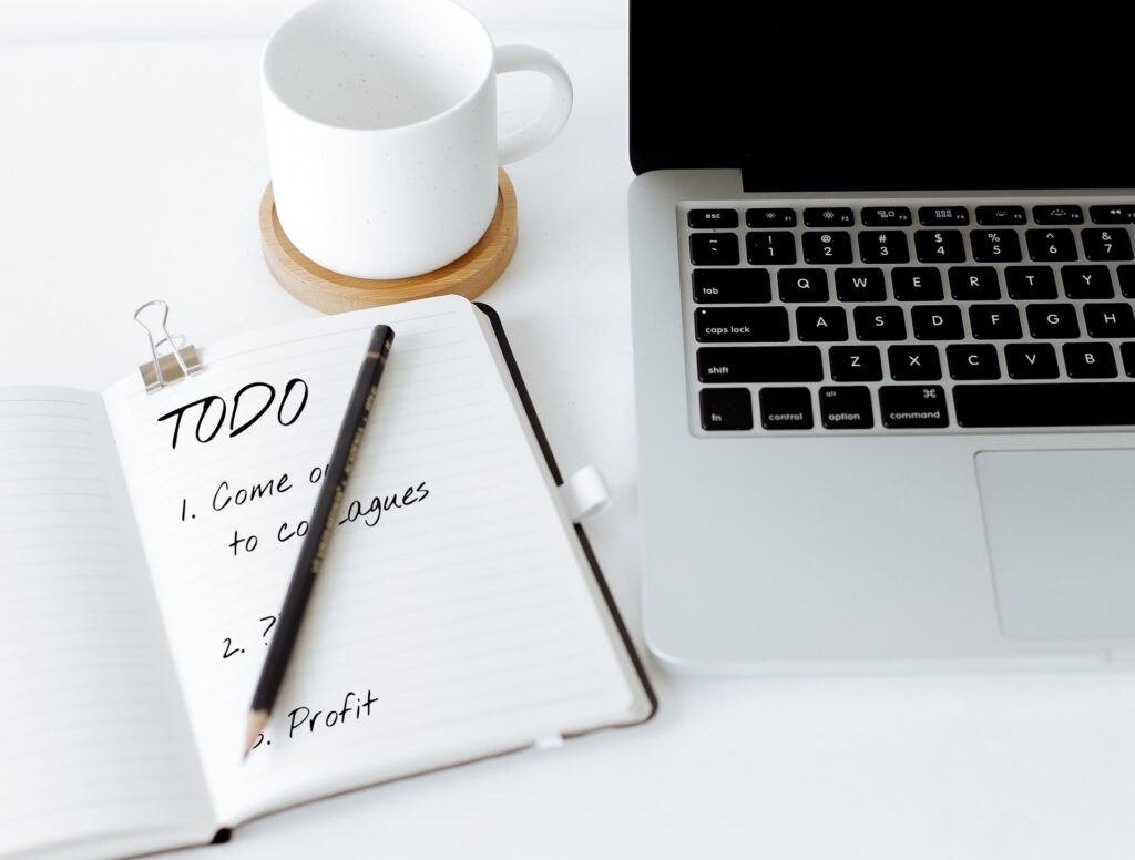 Laptop and mug alongside a notebook and pencil. In the notebook is written "TODO: 1. Come out to colleagues. 2. ???. 3. Profit."