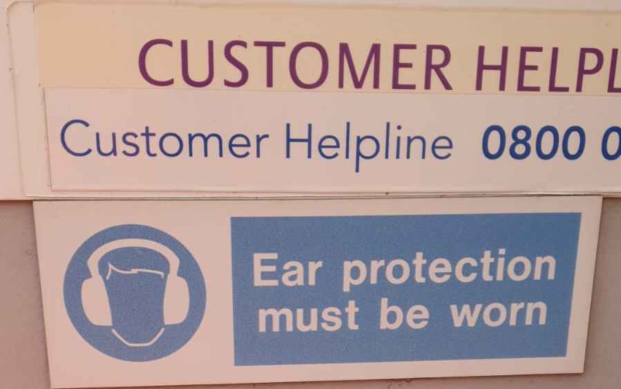 A sign advertising a number for a "Customer Helpline" directly above a sign that advises "Ear protection must be worn".