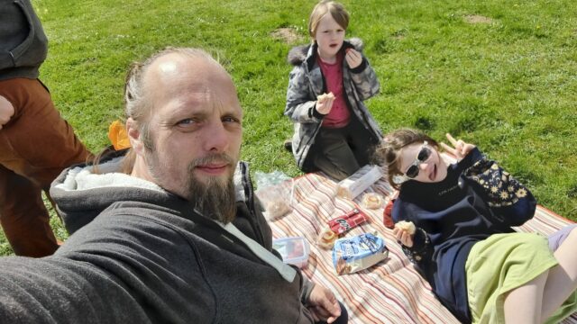 Dan kneels on a striped picnic mat with a 7-year-old and a 10-year old child alongside some sandwiches, iced fingers, Pepperami, fruit, and pretzels.
