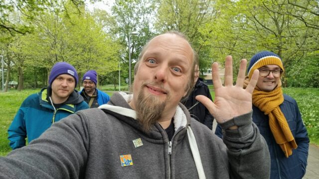 Dan waves to the camera; several other geeky folks walk along behind him in a leafy green city space.