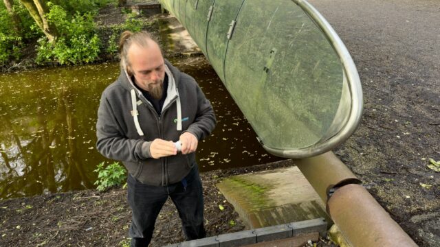 Dan on the bank of a water course, beneath a bridge, unrolls a logbook to sign his name.