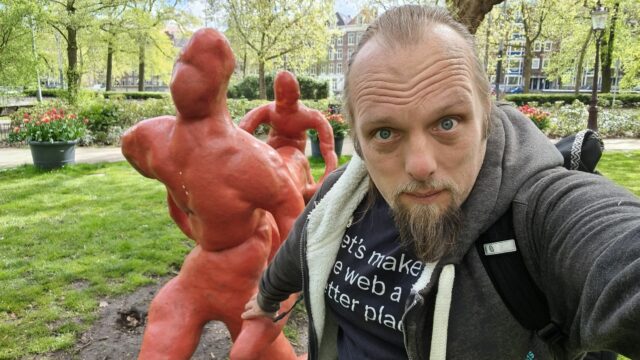 Dan holds the hand of a statue, as if guiding it, as it "carries" a log through a park.