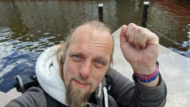 Dan sits on a pink triangular dock, holding a fist in the air. On his wrist can be sen the colours of the "bi pride" flag.