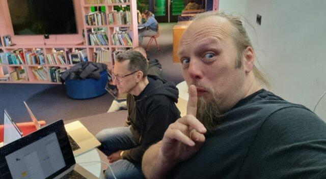 Dan, his finger to his lips and his laptop on his knees, makes a "shush" action. A coworker can be seen working behind him.