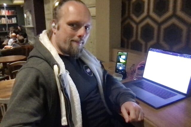 Dan sits at his laptop in a cafe; his phone is playing music to his earbuds.