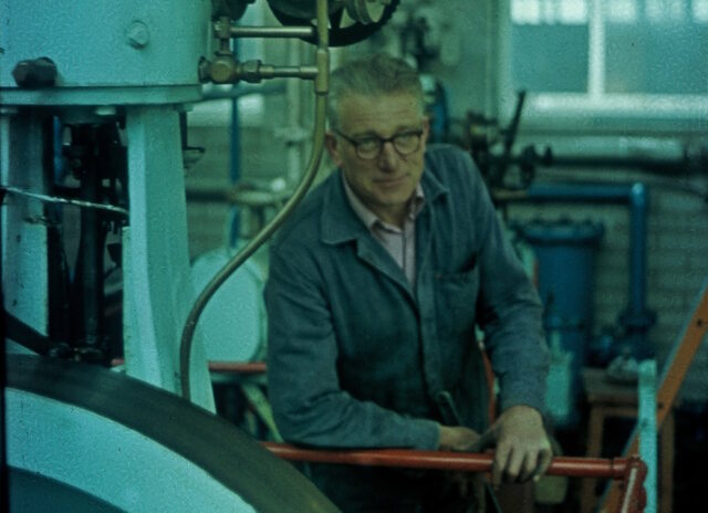 A grey-haired white man wearing spectacles and a boiler suit leans comfortably on a railing alongside industrial machinery.