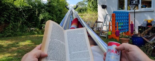 First-person view down a hammock in a rural garden. The hammock's occupier (Dan) is holding a novel and a can of premixed Pimms & lemonade.