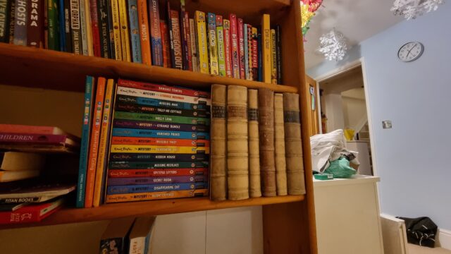 A bookcase in a child's bedroom, with Christmas decorations visible in the background. Several very old looking books stand conspicuously on the shelf.