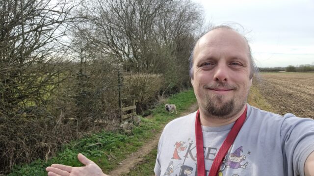 Dan, a red leash hung across his shoulders, gives a half-shrug as he smiles for a selfie on a grassy trail between a hedgerow and a field. Behind him, near a weathered old-fashioned footpath sign, his French Bulldog looks at him expectantly.