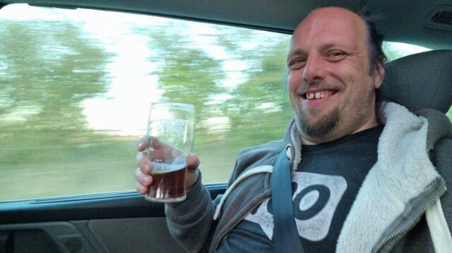 Dan, grinning, holding a quarter pint of beer in few rear passenger seat of a moving car.