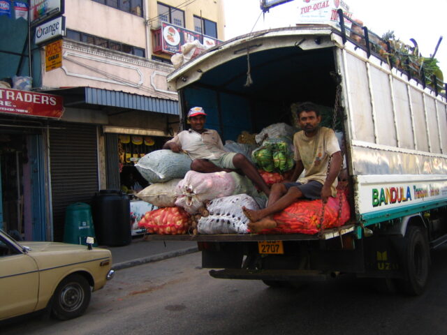 Two men sit in the back of an open-backed vegetable delivery truck.
