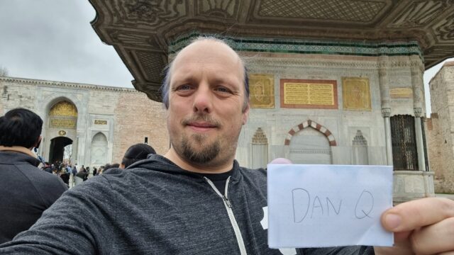 Dan holds up a sign showing his name in front of a gilded fountain decorated in Arabic script.