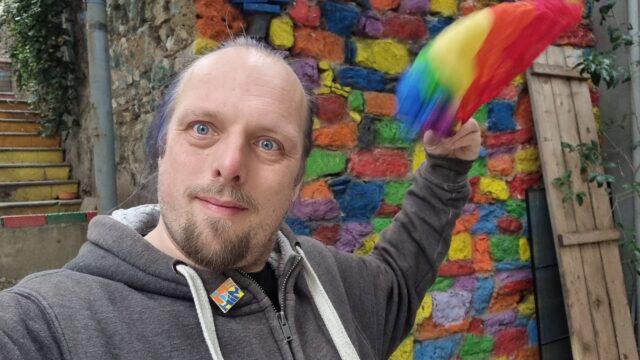 Dan waves a rainbow bandana in front of a wall painted in multitudinous colours, on a staircase with each step painted a different colour.