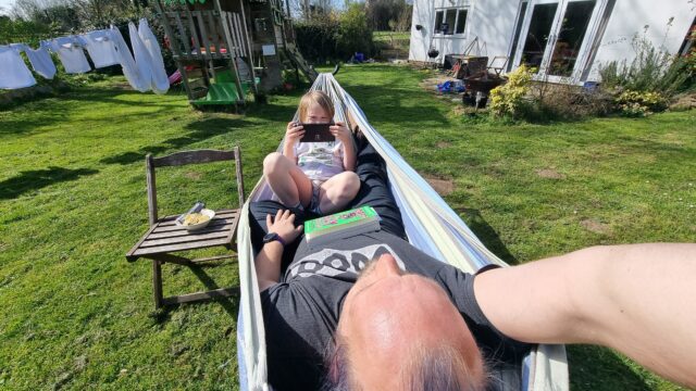 Dan, seen from his 'head' end, lies in a hammock with a green book, 'Bored Gay Werewolf' on his belly. At the other end of the hammock a boy plays on a Nintendo Switch. Around them is a garden containing a climbing frame and a washing line full of white shirts and sheets.