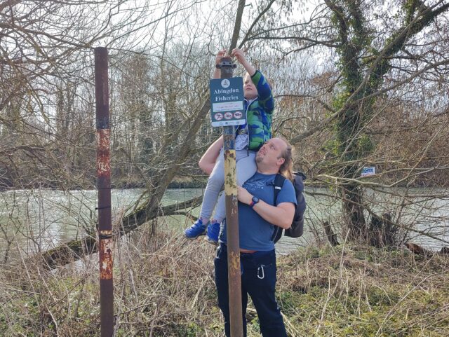 Dan lifts a child up to reach into the top of a post, with a sign detailing fishing regulations, alongside a lightly-wooded riverbank.