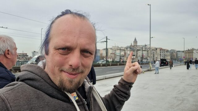 Dan stands on a busy, wide foot/road bridge, pointing at the top floor of a building overlooking the river that it crosses.