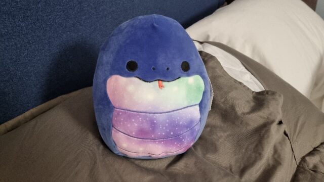 'Squishmallow' soft toy in the shape of a fat snake with a starry belly, sitting on a grey pillow.