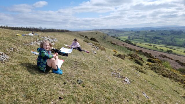 Two children on a rocky green hillside each draw on a sheet of A1 paper.