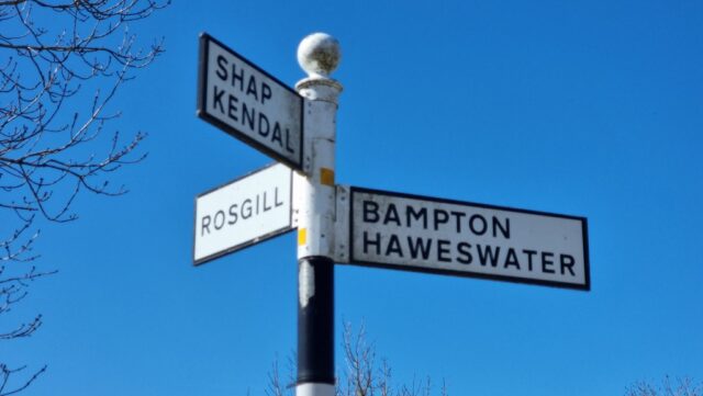 Pre-Worboys Committee British junction signpost in black and white, photographed against a bright blue sky. The signs point to Rosgill in one direction, Shap and Kendal in a second, and Bampton and Haweswater in a third.
