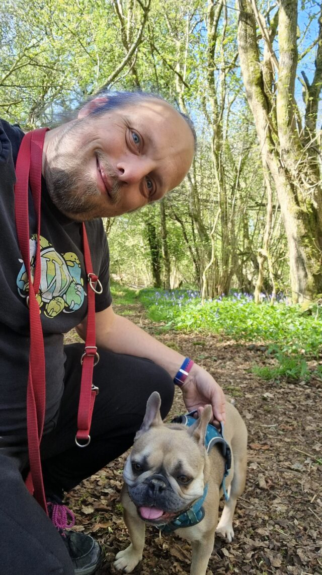 Dan, a dog lead hanging around his neck, crouches alongside his French Bulldog on a trail in a deciduous forest.