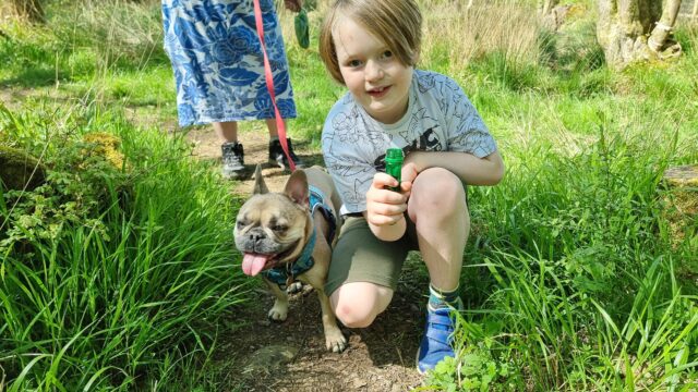 An 8-year-old boy in a white t-shirt and shorts crouches on a grassy trail, alongside a panting French Bulldog. The boy is holding a large green test tube.