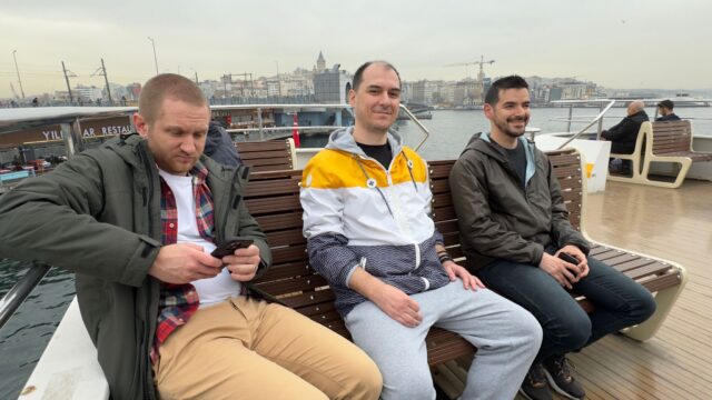 Three men sit on the upper deck of a passenger ferry near a bridge. A city skyline can be seen in the background.