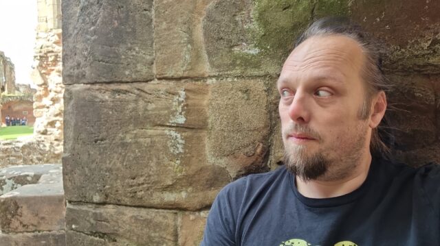 Dan, looking shifty, against a red stone castle wall.
