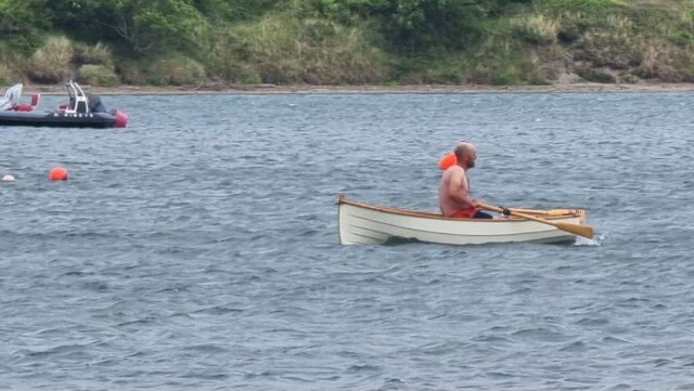 A shirtless white bald man rows a small white boat past bouys in a choppy estuary.