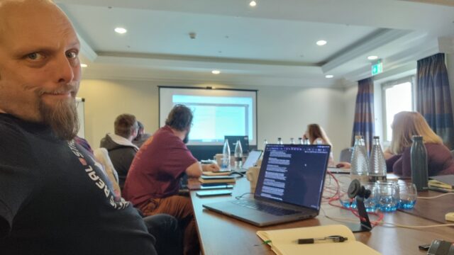 Dan sits at his laptop in front of a long conference table where a group of people are looking at a projector screen.