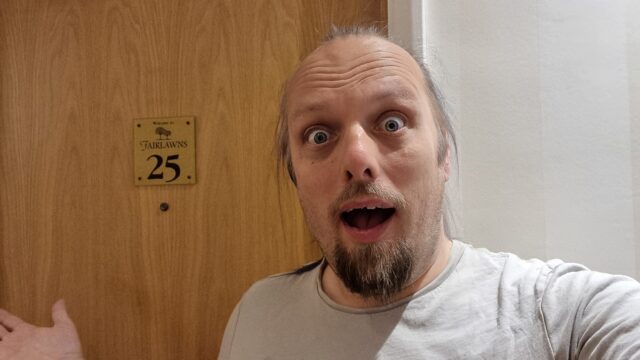 Dan looks shocked as he stands in front of a wooden door on which a brass plaque reads 'Welcome to Fairlawns 25'.