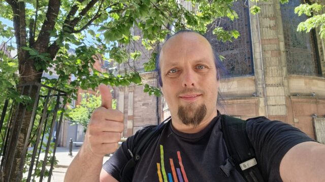 Under a shady tree and in front of a church, Dan throws a thumbs-up.
