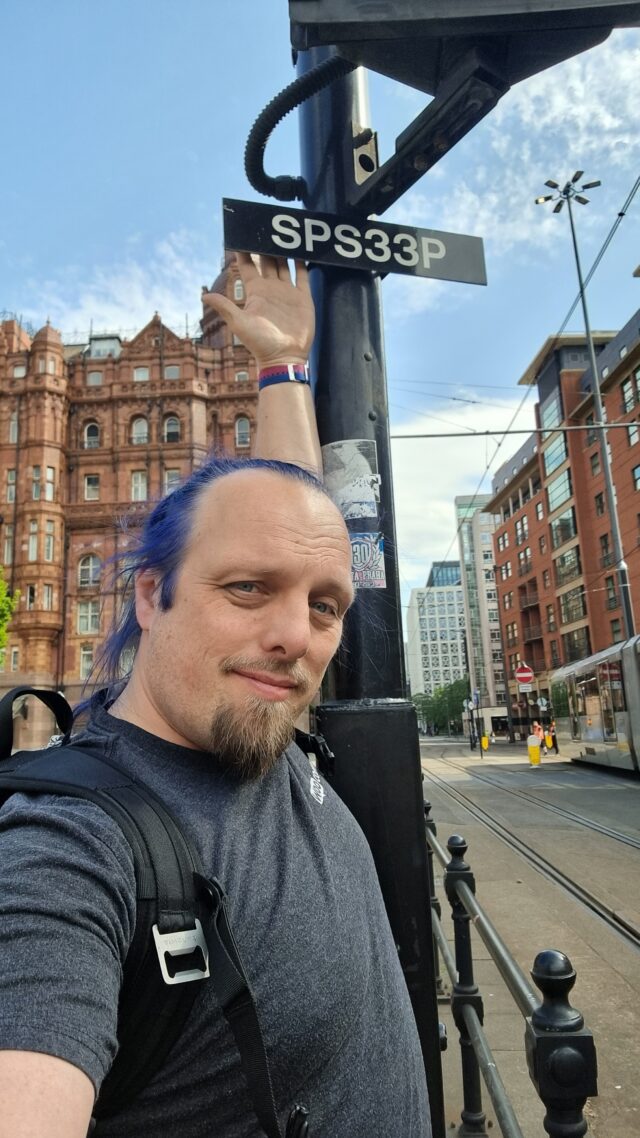 Dan stretches high to touch the back of a sign mounted on a pole, alongside tram signals, in a city centre.