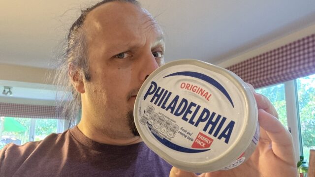 Dan, a white man with blue hair and a goatee, wearing a purple t-shirt, stands in a spacious residential kitchen holding a 'Family Pack'-sized tub of 'Original Philadelphia'.