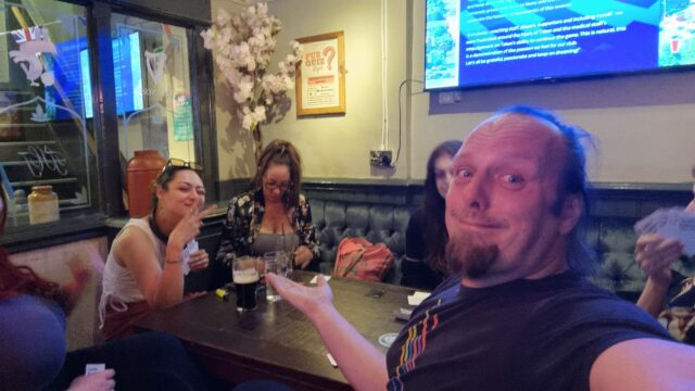 Dan shrugs for the camera as he sits at a pub table with a variety of people; one woman, sat to his left, throws a V-sign with her fingers.